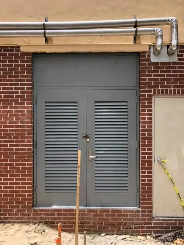 Grey louvered double doors under a transom in a red brick wall